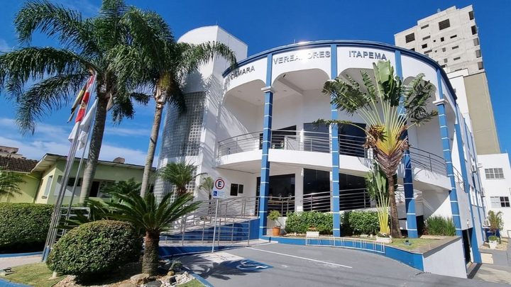 Fachada da Câmara de Vereadores de Itapema, prédio branco com colunas azuis e o nome da instituição em letras na parte alta, com palmeiras e mastros de bandeira em frente.
