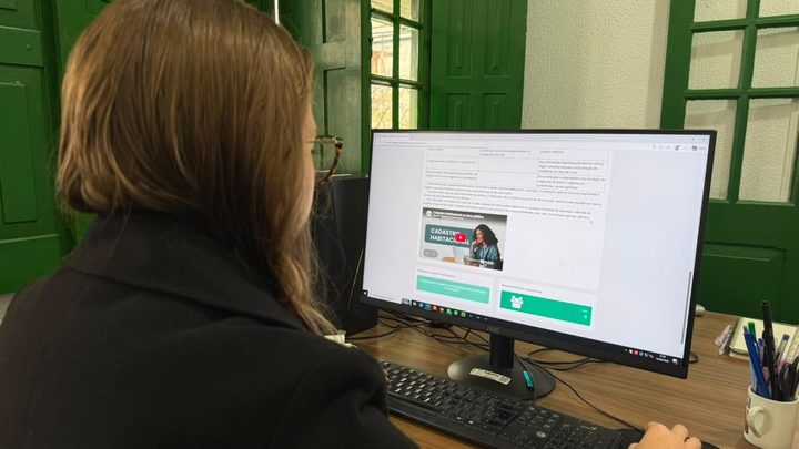 Pessoa vista de costas diante de um monitor que exibe a página da Prefeitura com o banner do cadastro habitacional, sobre mesa de madeira com teclado e mouse.