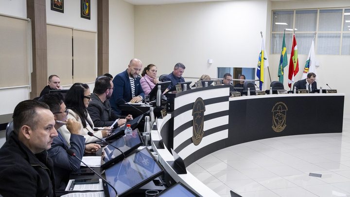 Vereadores durante sessão no plenário da Câmara de Itapema, sentados na bancada com microfones e monitores à frente.