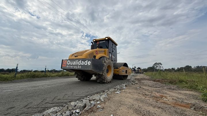 Rua em obras com maquinário sobre o leito de terra preparado para pavimentação, meio-fio novo assentado e casas ao longo da via.