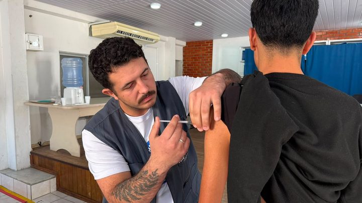 Profissional de saúde aplicando vacina no braço de uma pessoa sentada em unidade de saúde, com bandeja de insumos ao lado.