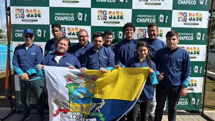 Delegação paradesportiva de Itapema posando com a bandeira do município diante de um painel do Parajasc e da Prefeitura de Chapecó.