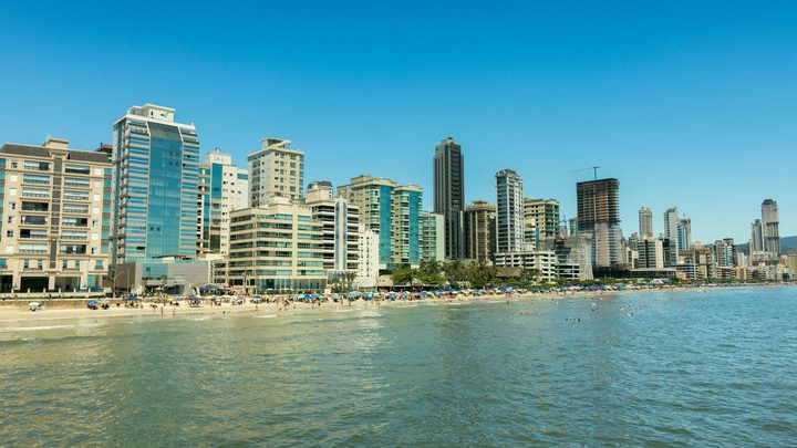 Orla verticalizada de Itapema vista do mar, com faixa de areia estreita ocupada por guarda-sóis coloridos e uma parede de edifícios altos logo atrás.