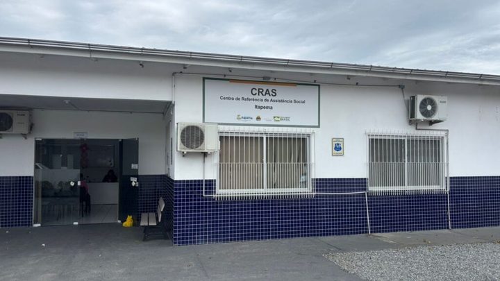 Fachada do Centro de Referência de Assistência Social de Itapema, prédio branco com faixa de azulejo azul, placa do CRAS acima da entrada e pátio de concreto à frente.