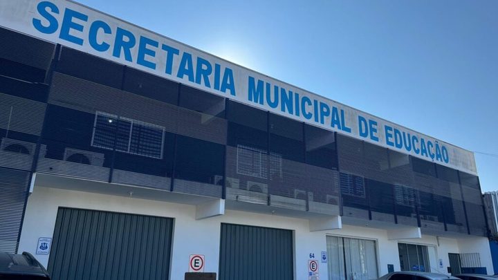 Fachada da Secretaria Municipal de Educação de Itapema, prédio de dois andares com o nome em letras azuis no alto, grades escuras no pavimento superior e portões verdes na frente.