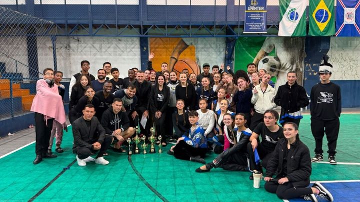 Integrantes da Banda Municipal de Itapema reunidos em quadra coberta para foto de grupo, com os troféus conquistados apoiados no chão à frente e bandeiras penduradas ao fundo.