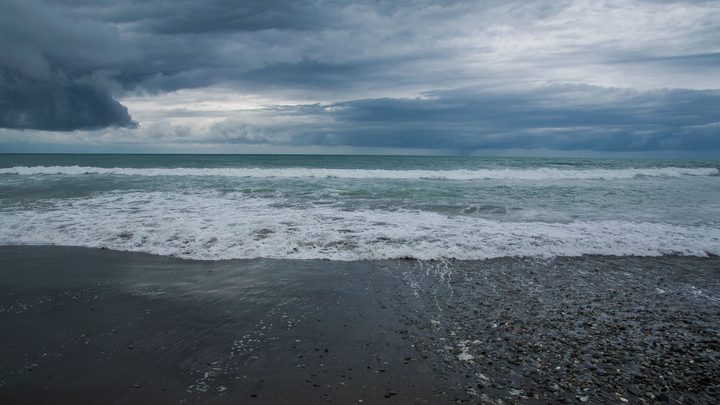 Nuvens carregadas e escuras de temporal ocupando todo o céu sobre o mar, com a arrebentação branca avançando sobre a areia escura da praia.