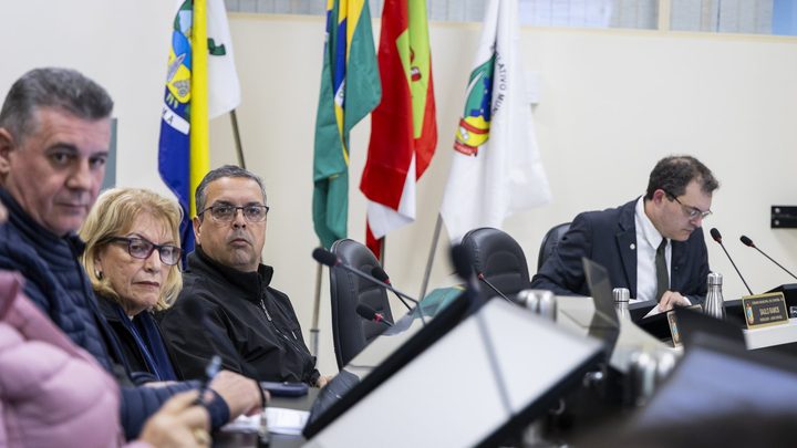 Vereadores sentados à mesa do plenário da Câmara de Itapema, com microfones à frente e as bandeiras do município, do estado e do Brasil ao fundo.