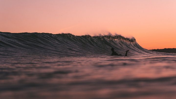 Surfista deitado na prancha esperando a onda quebrar ao entardecer, em silhueta contra o céu alaranjado, com a parede da onda se formando ao lado.