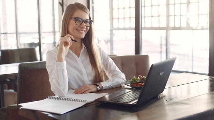 Mulher de camisa branca e óculos sentada a uma mesa de madeira, sorrindo com uma caneta na mão, ao lado de um caderno aberto e de um notebook ligado.