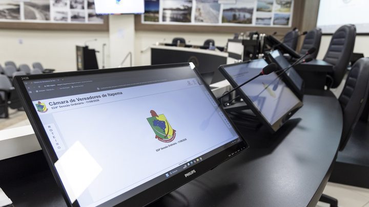 Plenário vazio da Câmara de Itapema visto de perto da bancada, com dois monitores ligados sobre a mesa curva, microfones de haste e cadeiras pretas.
