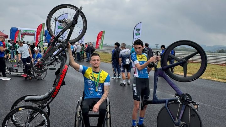 Dois atletas de Itapema no asfalto molhado de um autódromo. À esquerda, um paraciclista de cadeira de rodas e medalha no peito ergue a handbike com a mão. À direita, um ciclista segura uma bicicleta de contrarrelógio de rodas fechadas.
