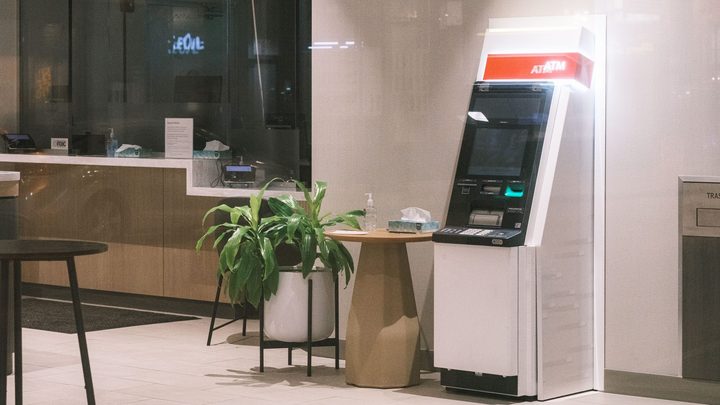 Saguão de agência bancária com um caixa eletrônico branco encostado na parede, uma planta em vaso ao lado e o balcão de atendimento vazio ao fundo.