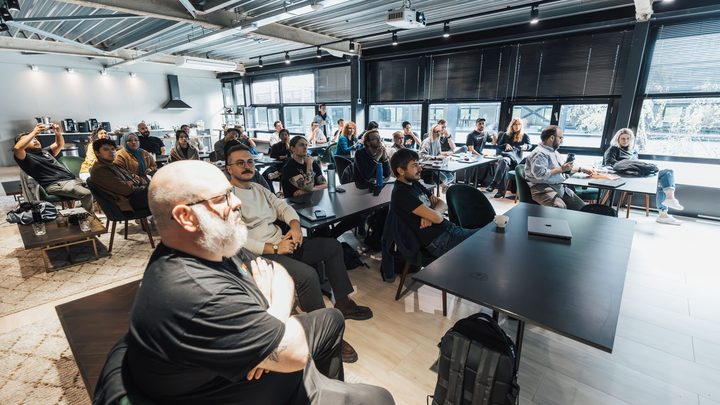 Sala de reunião cheia de adultos sentados em cadeiras e em volta de mesas escuras, todos olhando na mesma direção, com janelas grandes ao fundo.