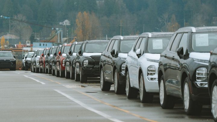 Fileira de carros e utilitários novos estacionados lado a lado num pátio de concreto molhado, com etiquetas coladas nos vidros.