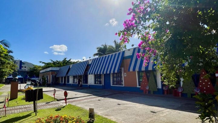 Prédio térreo de Itapema com toldos listrados de azul e branco sobre as vitrines, calçadão de concreto na frente e canteiro de flores amarelas e laranjas na grama.