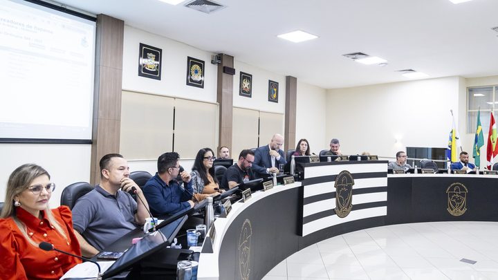 Plenário da Câmara de Itapema durante sessão, com vereadores sentados na bancada curva preta e branca diante de microfones e monitores, e bandeiras ao fundo.