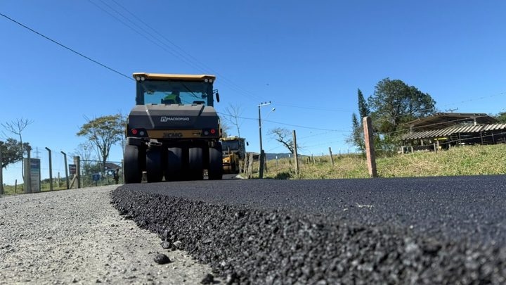 Rolo compactador amarelo e preto sobre uma faixa de asfalto novo em estrada rural, visto rente ao chão, com cerca, potreiro e galpão de telhado baixo ao lado.