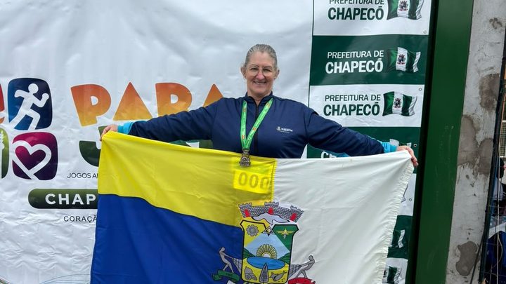 Atleta de Itapema com uniforme azul e medalha de prata no peito, sorrindo enquanto abre a bandeira do município diante de um painel do Parajasc.