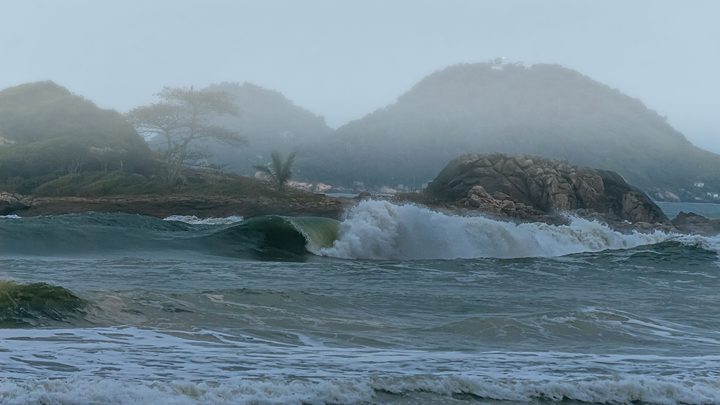 Onda grande quebrando perto da areia num dia de neblina, com espuma branca subindo à frente de um costão de pedras e morros verdes encobertos pela bruma ao fundo.