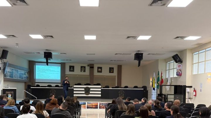 Plenário claro com fileiras de cadeiras pretas ocupadas por servidores vistos de costas, um palestrante de terno em pé ao microfone diante da mesa diretora e um telão com slide de texto projetado na parede.