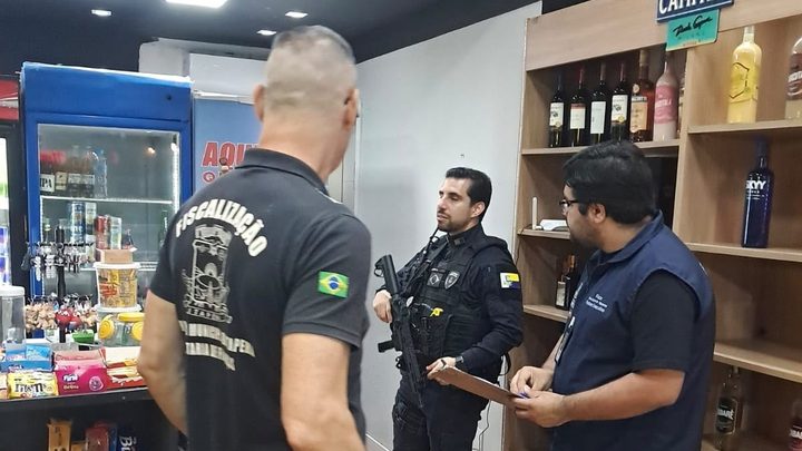 Três agentes de pé dentro de uma conveniência, um deles de costas com a camisa escrita Fiscalização Prefeitura Municipal de Itapema, um guarda municipal fardado de preto no centro e um fiscal com prancheta à direita, diante de prateleiras com garrafas.
