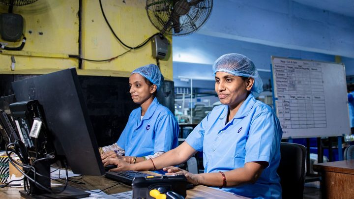 Duas trabalhadoras de uniforme azul e touca descartável sentadas lado a lado diante de computadores numa mesa de trabalho de fábrica, com um quadro branco de metas na parede ao fundo.