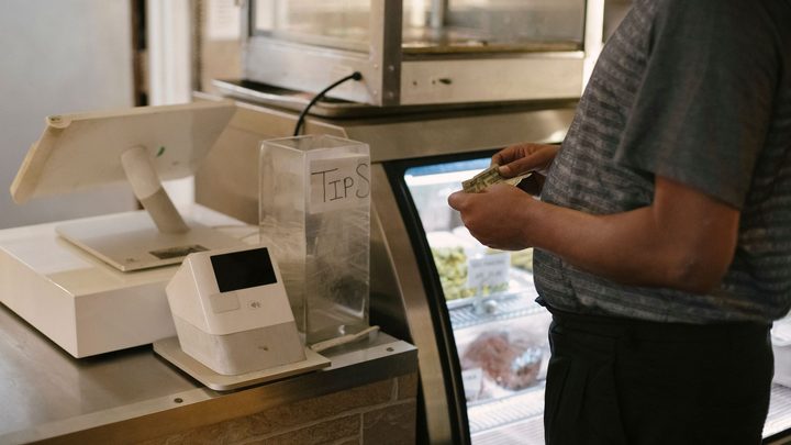 Balcão de um comércio pequeno com máquina de cartão e caixa registradora brancos sobre a bancada de inox, ao lado de uma vitrine refrigerada, e uma pessoa de camiseta cinza segurando dinheiro, sem que o rosto apareça.