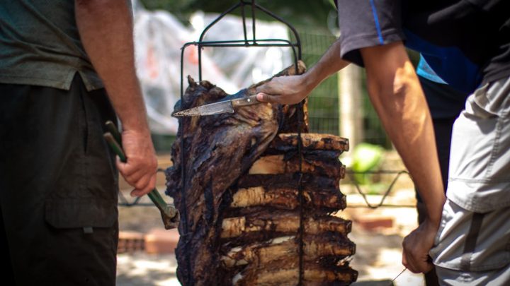 Costela assada na vertical numa grelha de ferro sobre brasas ao ar livre, com dois homens em volta, um segurando a faca e outro o espeto, sem que os rostos apareçam.