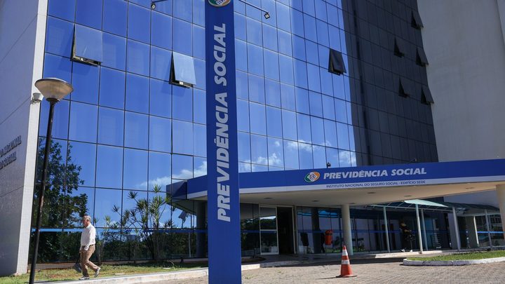 Fachada envidraçada do edifício sede do INSS em Brasília, com um totem azul alto escrito Previdência Social na frente e o céu refletido nos vidros.