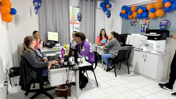 Sala de atendimento com mesas brancas, computadores e impressoras, servidoras atendendo pessoas sentadas em cadeiras pretas, com balões azuis e laranja decorando as paredes.