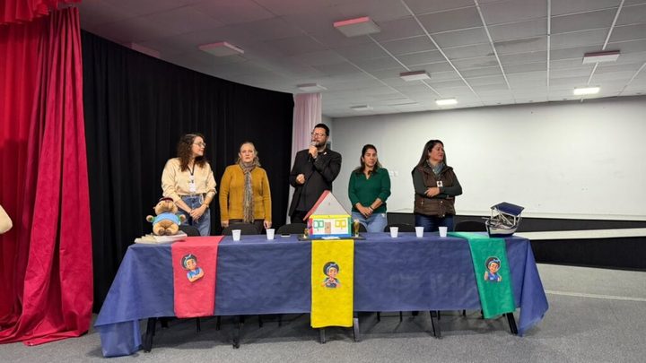 Cinco pessoas de pé atrás de uma mesa comprida coberta por toalha azul, num auditório de cortina vermelha, com um homem de terno falando ao microfone e uma maquete de casa colorida sobre a mesa.