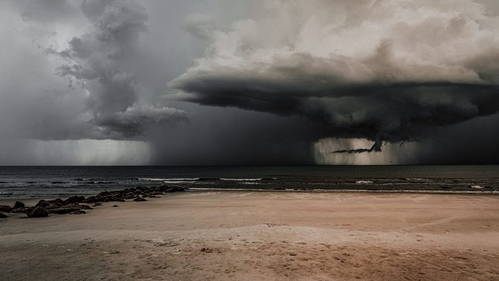 Praia deserta de areia clara com pedras na margem esquerda e uma nuvem escura de tempestade despejando chuva sobre o mar ao fundo, sob céu carregado.