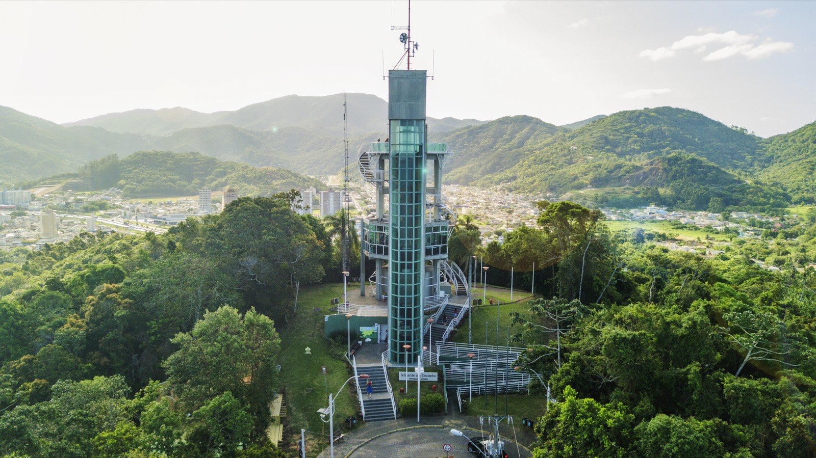 Torre mirante de vidro cercada de mata fechada, com morros verdes ao fundo e a área urbana espalhada pelo vale ao redor.