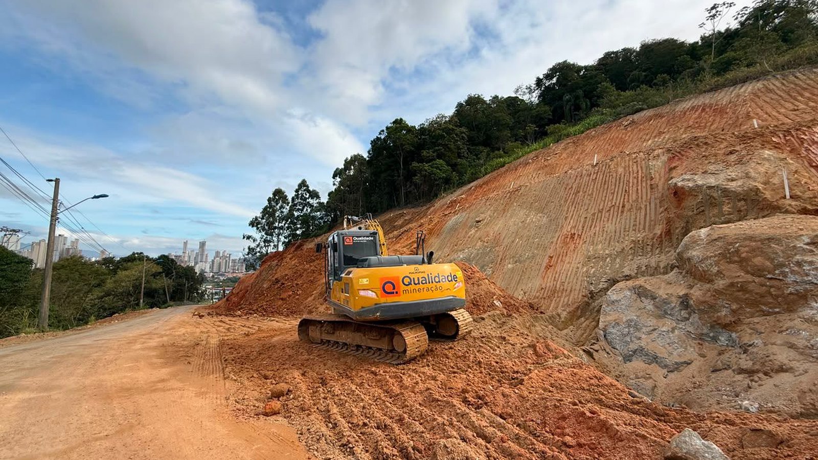 Escavadeira amarela trabalhando sobre uma via de terra no Morro do Encano, em Itapema, ao lado de um talude de encosta recém-cortado com paredão de rocha exposta, sob céu de nuvens e com prédios altos ao longe.