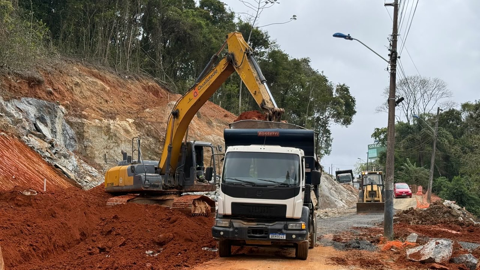 Escavadeira amarela com o braço estendido despejando terra vermelha na caçamba de um caminhão branco parado numa estrada de terra do Morro do Encano, em Itapema, com o barranco recém-cortado e paredão de rocha exposta à esquerda, poste de energia à direita e outro caminhão e uma pá carregadeira ao fundo.