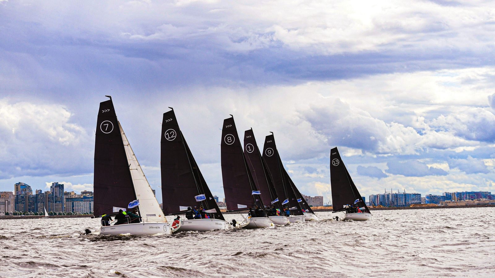 Sete veleiros pequenos de vela escura e numerada largando lado a lado em mar encrespado, com as tripulações debruçadas na borda e a linha de prédios da cidade ao fundo, sob céu de nuvens carregadas.