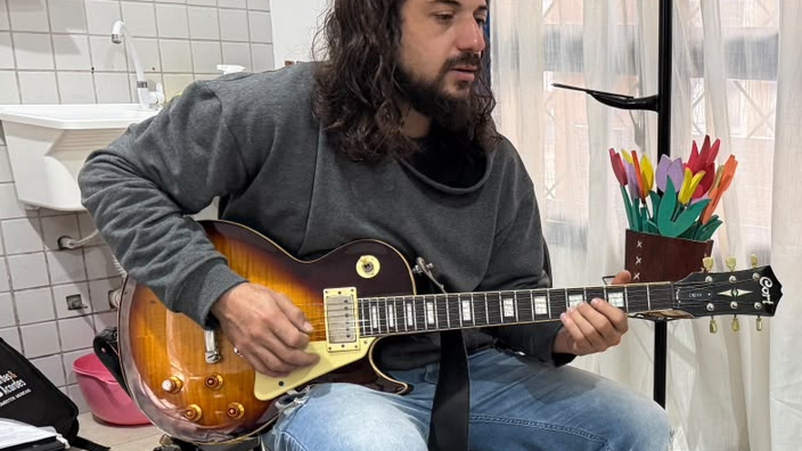 Instrutor sentado em banqueta tocando guitarra numa sala de oficina, com amplificador, cabos e suporte de partitura ao redor.