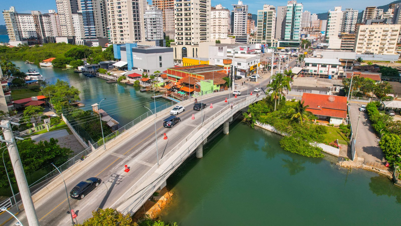 Vista aérea de Itapema com a ponte sobre o rio, carros passando entre cones laranja, casas e comércios de um lado, embarcações ancoradas do outro e a linha de prédios altos ao fundo.