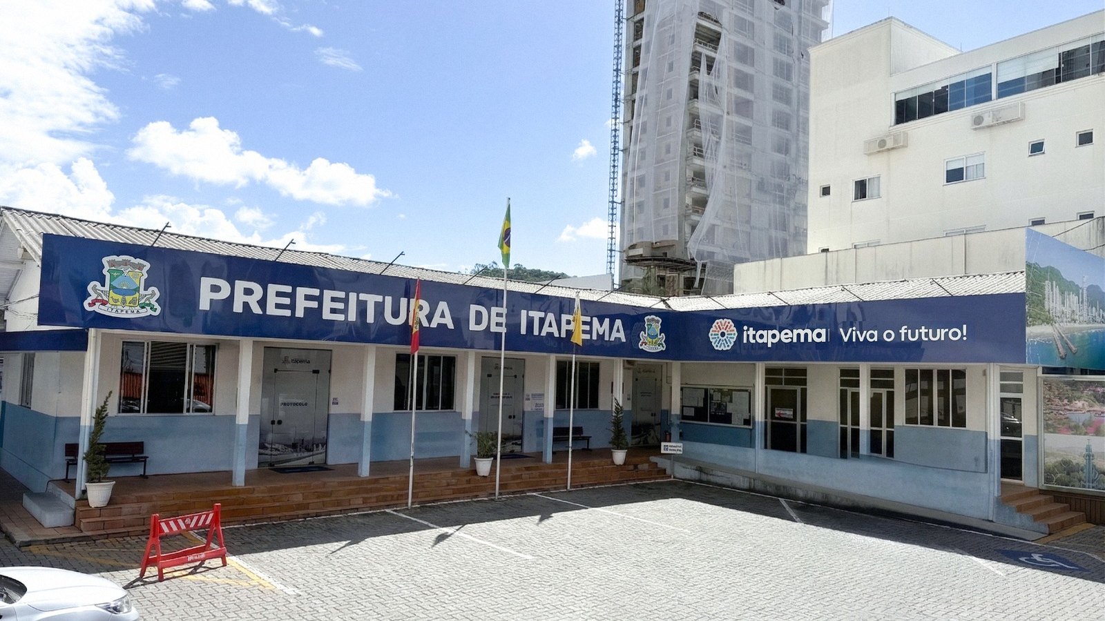 Prédio da Prefeitura de Itapema visto de fora, com fachada clara, janelas em fita e bandeiras hasteadas na entrada.