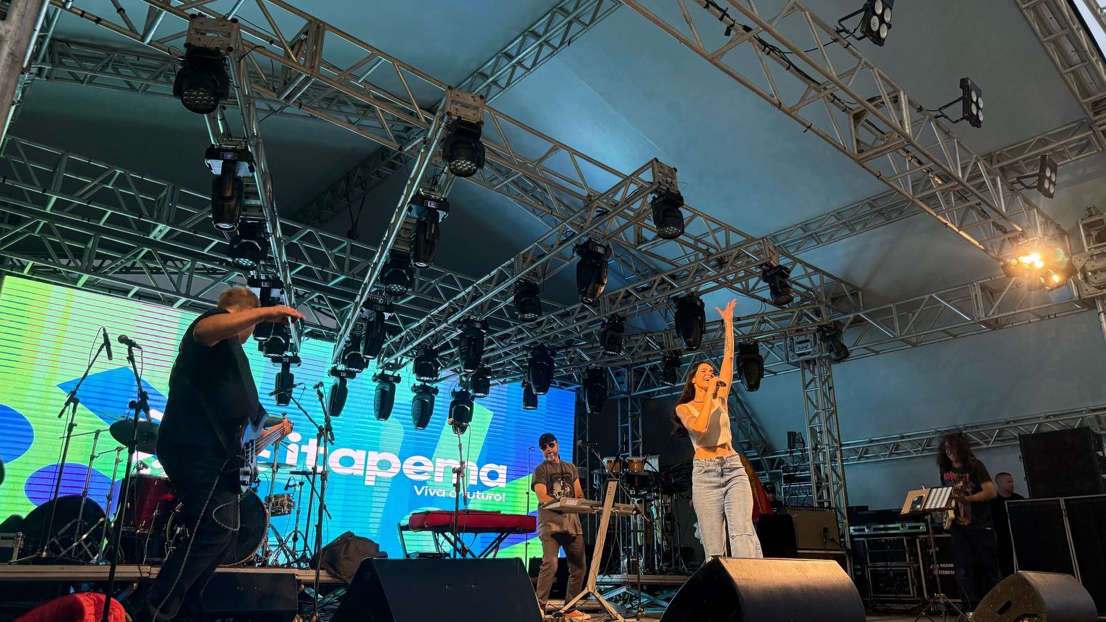 Banda tocando em palco coberto com estrutura de treliça e refletores, com um telão ao fundo exibindo a marca Itapema Viva o Futuro.