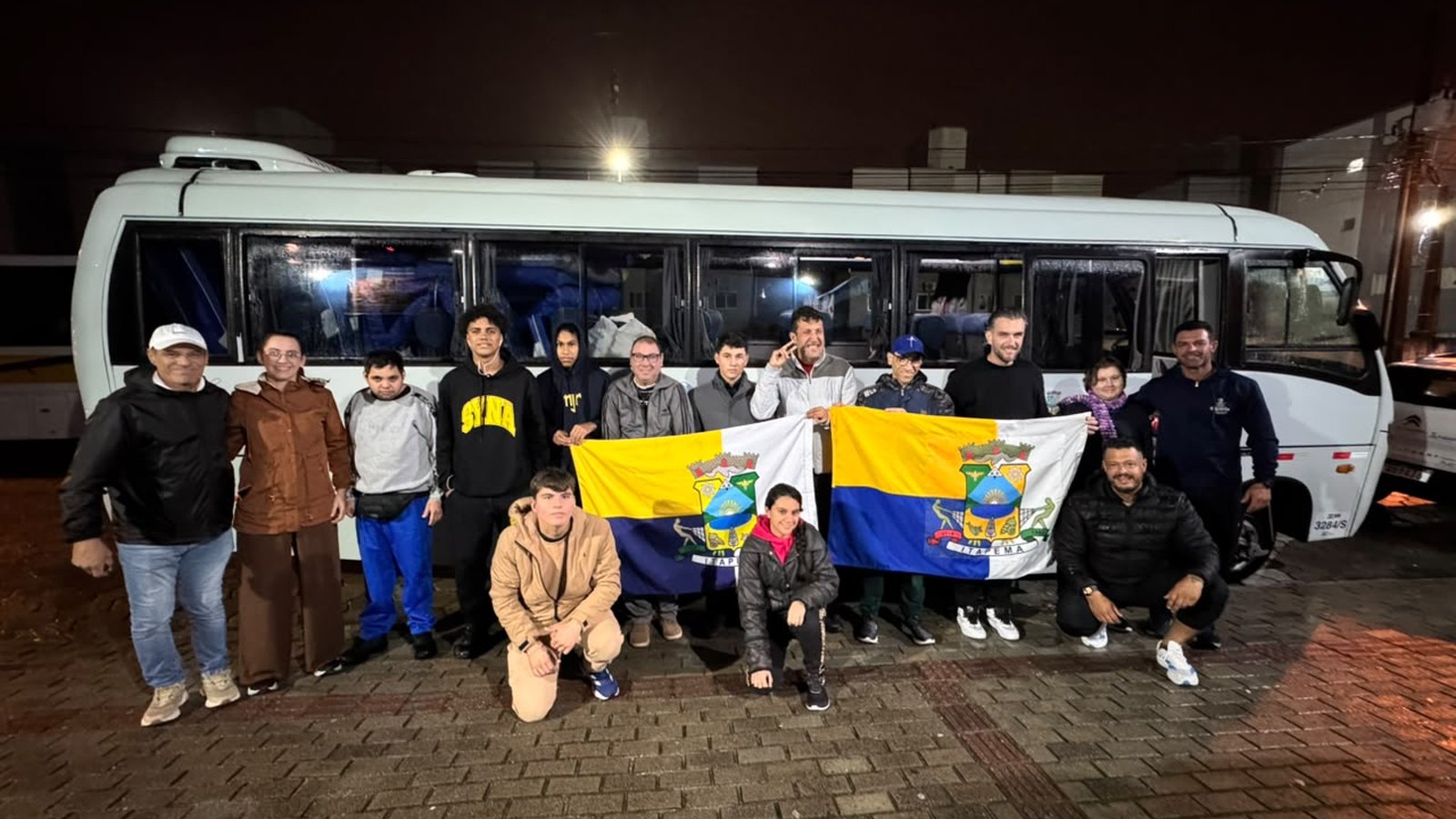 Delegação de atletas e técnicos posando à noite em frente a um ônibus branco, segurando duas bandeiras do município de Itapema.