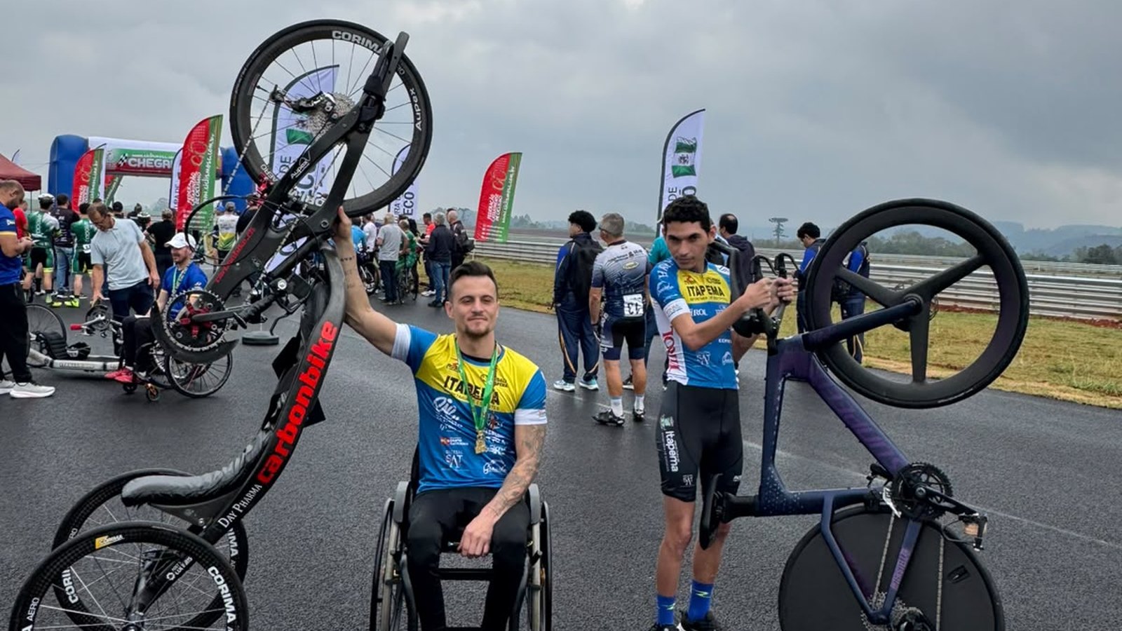 Dois atletas de Itapema no asfalto molhado de um autódromo, com bandeirolas e o pórtico de chegada ao fundo. À esquerda, um paraciclista de cadeira de rodas e medalha no peito ergue a handbike com a mão. À direita, um ciclista de uniforme azul segura uma bicicleta de contrarrelógio de rodas fechadas.