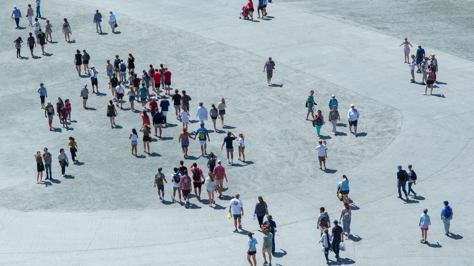 Vista de cima de uma praça de piso claro em dia de sol, com dezenas de pessoas caminhando espalhadas pelo chão, algumas em grupo e outras sozinhas, projetando sombras curtas.