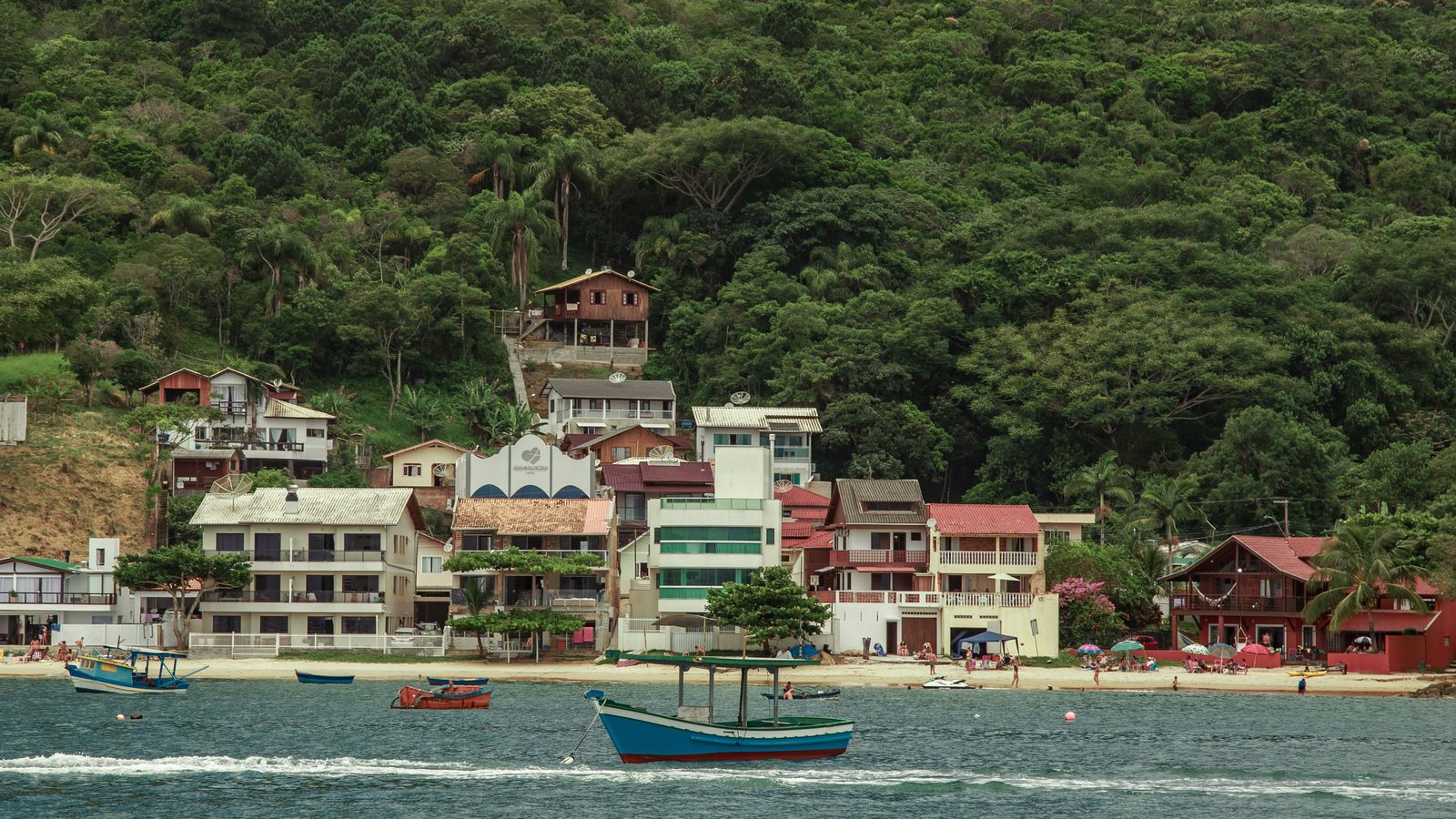 Casas coloridas na beira da água em Porto Belo, com barcos de pesca ancorados em primeiro plano e morros cobertos de mata ao fundo.