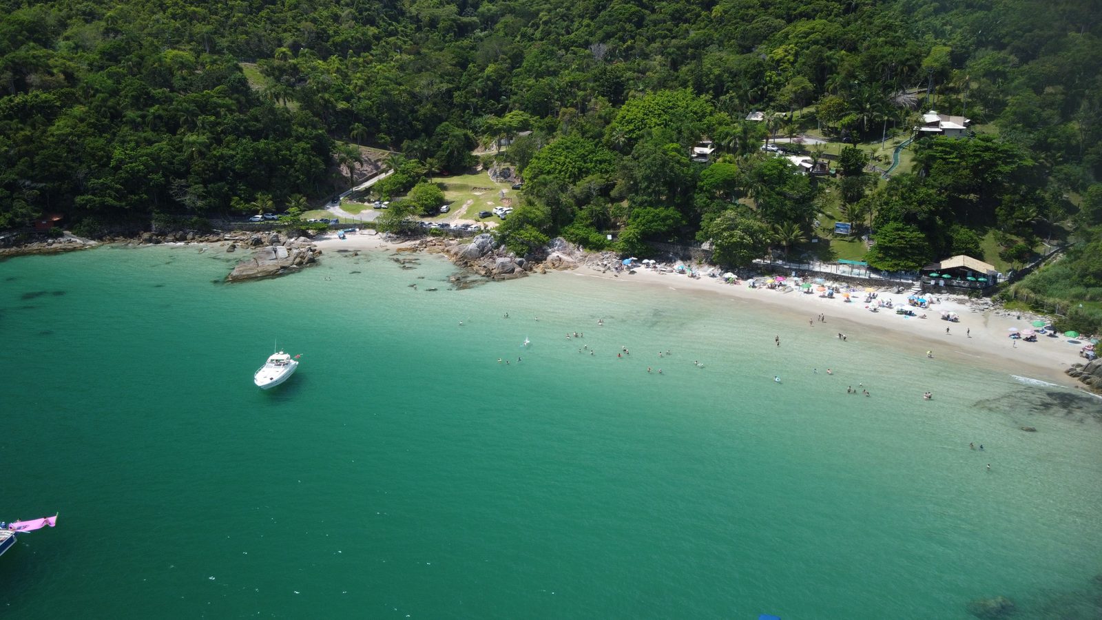 Vista aérea de uma enseada de Porto Belo: água esverdeada e transparente ocupa a metade inferior da imagem, uma lancha branca está ancorada e, na faixa de areia ao fundo, guarda-sóis se espalham diante de um morro coberto de mata fechada.