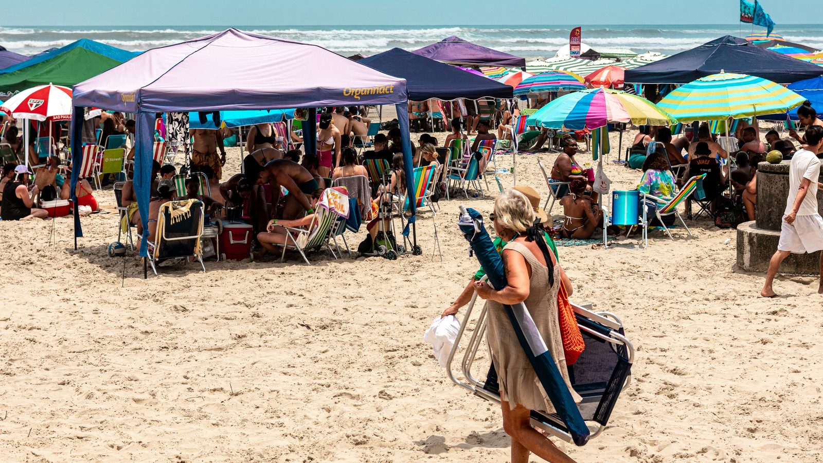 Praia cheia em dia de sol, com tendas e guarda-sóis coloridos ocupando a areia, cadeiras de praia espalhadas e o mar ao fundo.