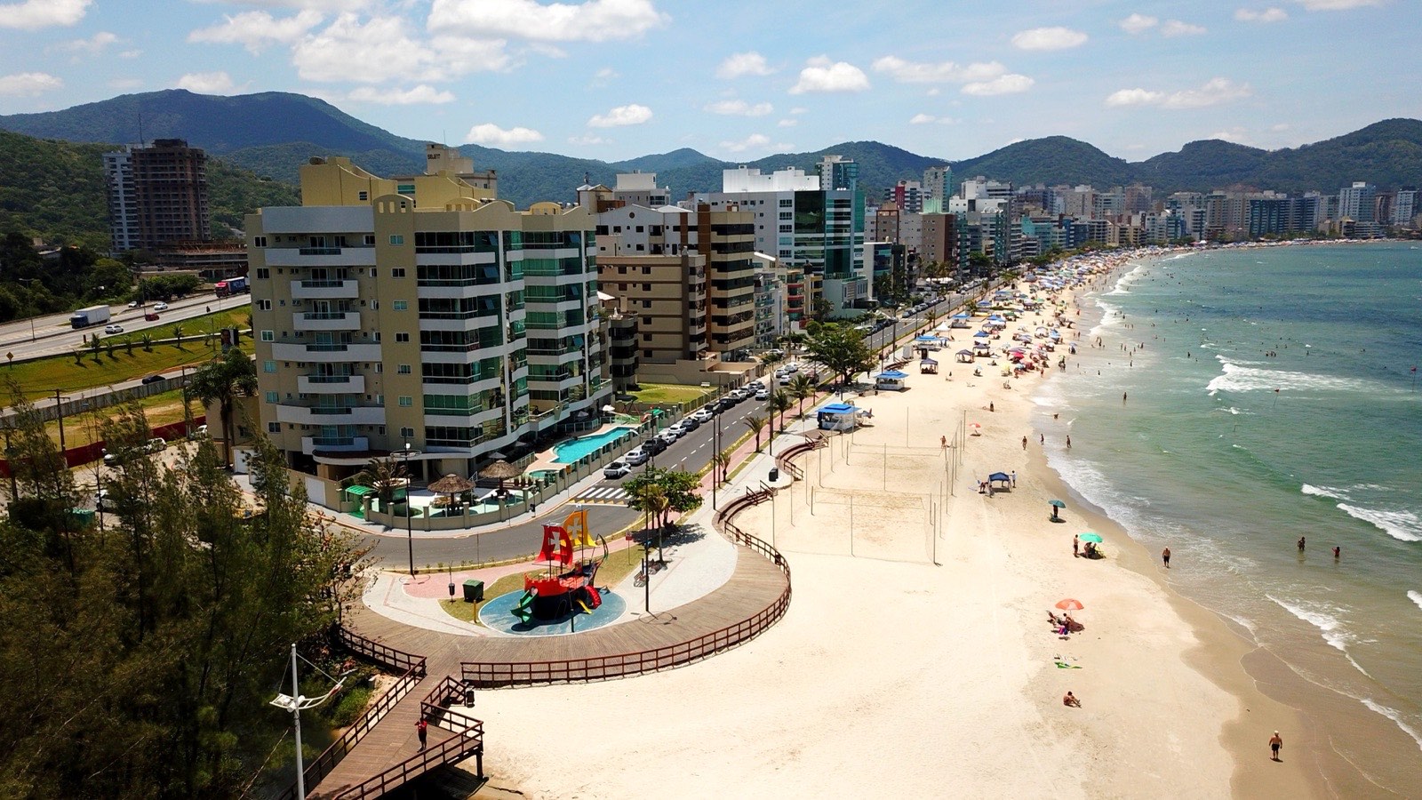 Vista aérea da praia do Centro de Itapema, com a avenida beira-mar, o calçadão, um parquinho infantil, prédios residenciais à esquerda e morros verdes ao fundo.