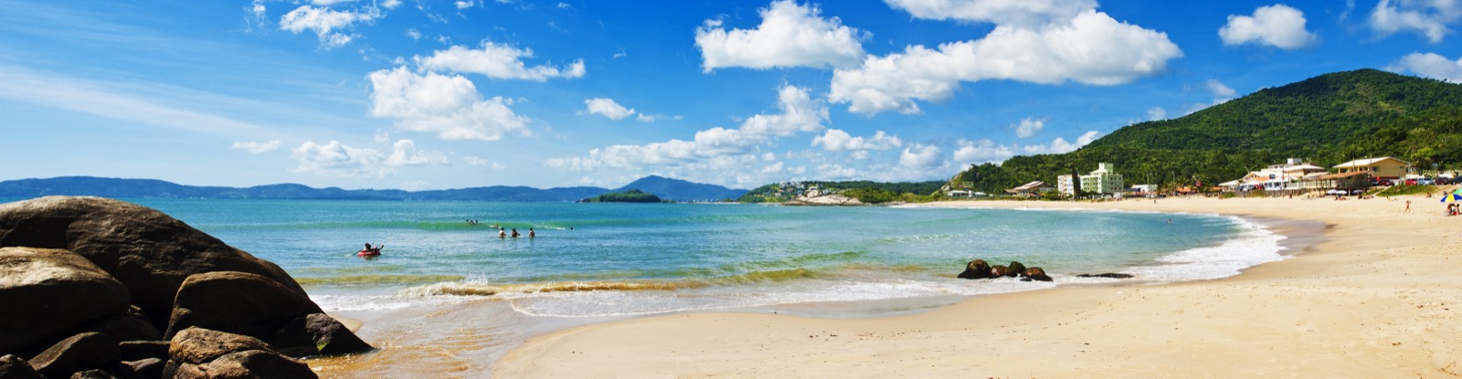 Panorâmica da praia do Ilhota, em Itapema: costões de pedra à esquerda, mar esverdeado ao centro, faixa de areia larga e o morro coberto de mata à direita.