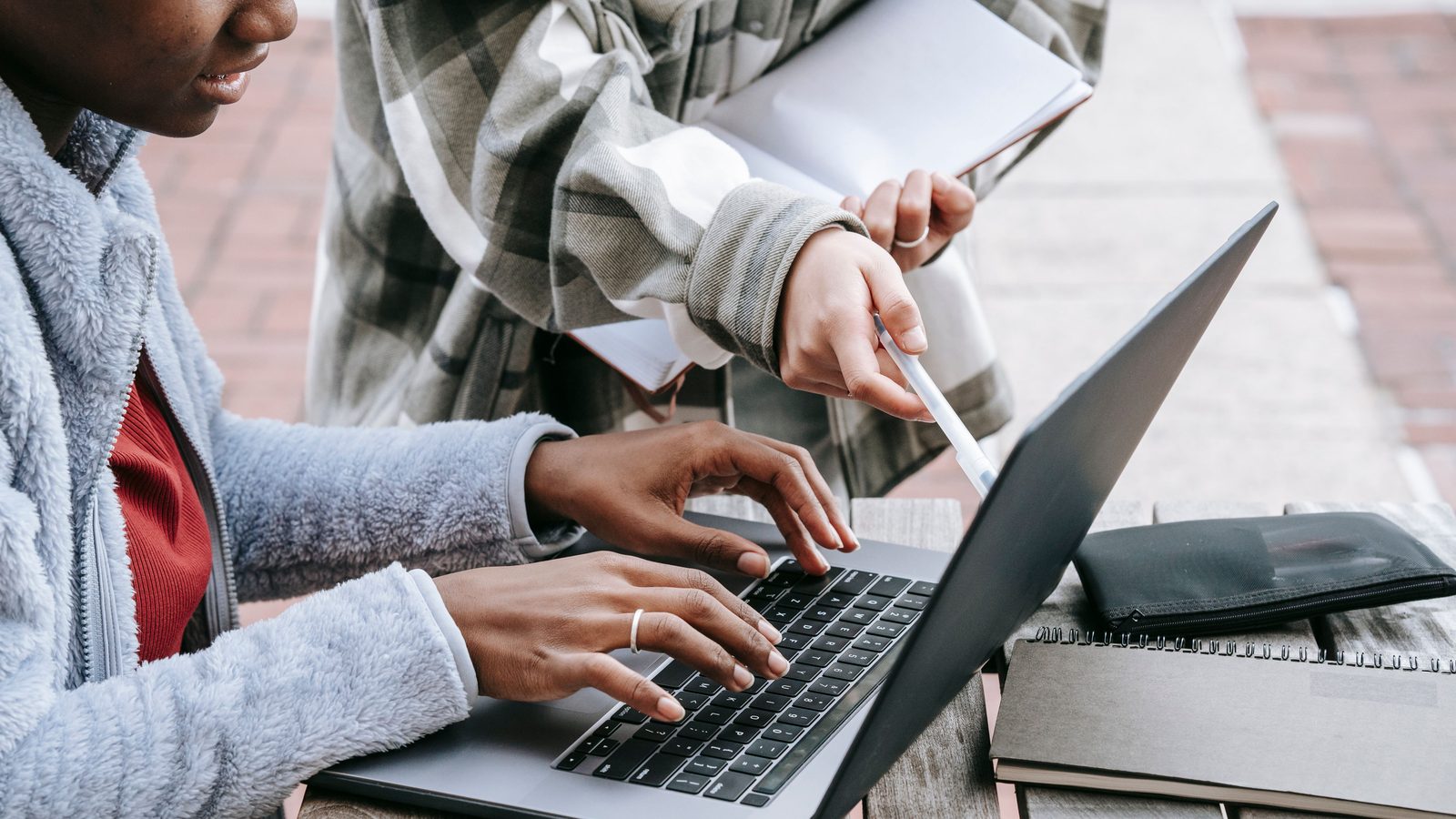 Duas estudantes sentadas num banco de praça digitando num notebook aberto sobre a mesa, com cadernos empilhados ao lado e uma delas apontando a tela com a caneta.